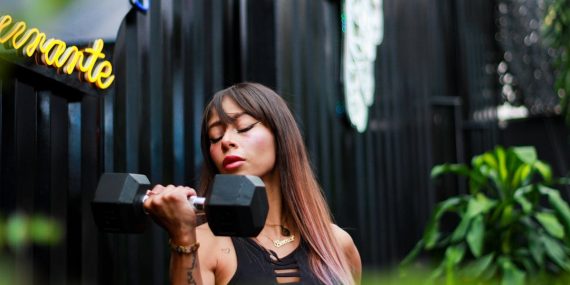 Fit woman with long hair and tattoos lifting a dumbbell with intense focus outdoors.